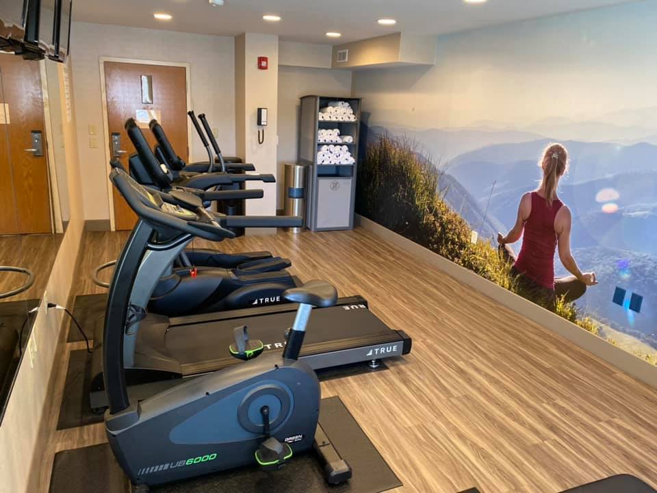 a picture of the fitness center at the newly renovated Clarion Pointe Harrisonburg featuring a wall mural of a woman meditating on a mountaintop, one treadmill, one elliptical, one exercise bike. 3 televisions, and more. 